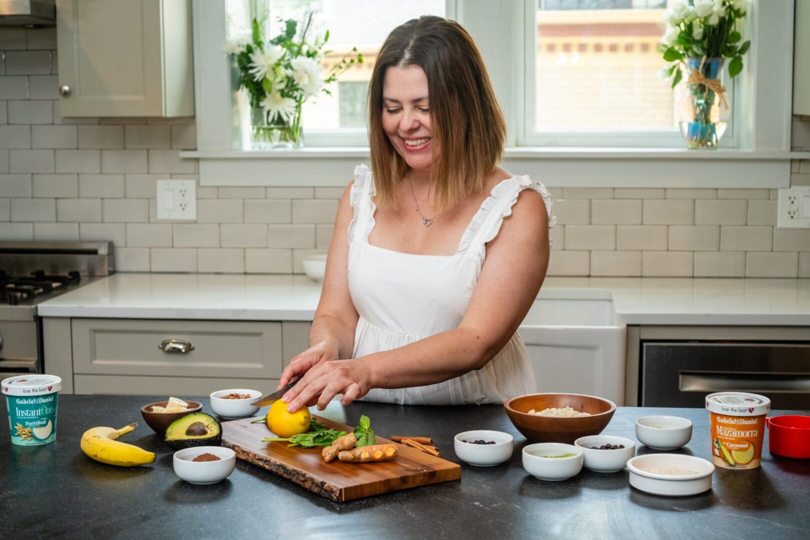 A woman in a white top smiles while slicing a lemon in a modern kitchen, surrounded by various ingredients and cooking utensils.