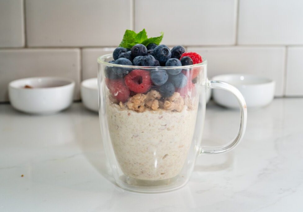 Glass cup of oatmeal topped with blueberries, raspberries, and a mint leaf on a white kitchen counter.